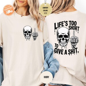 May include: Ivory-colored t-shirt featuring a black skull graphic with a middle finger gesture. The back of the shirt displays the text "LIFE'S TOO SHORT TO GIVE A SHIT," along with a skull and hand graphic. The shirt is a comfort color.