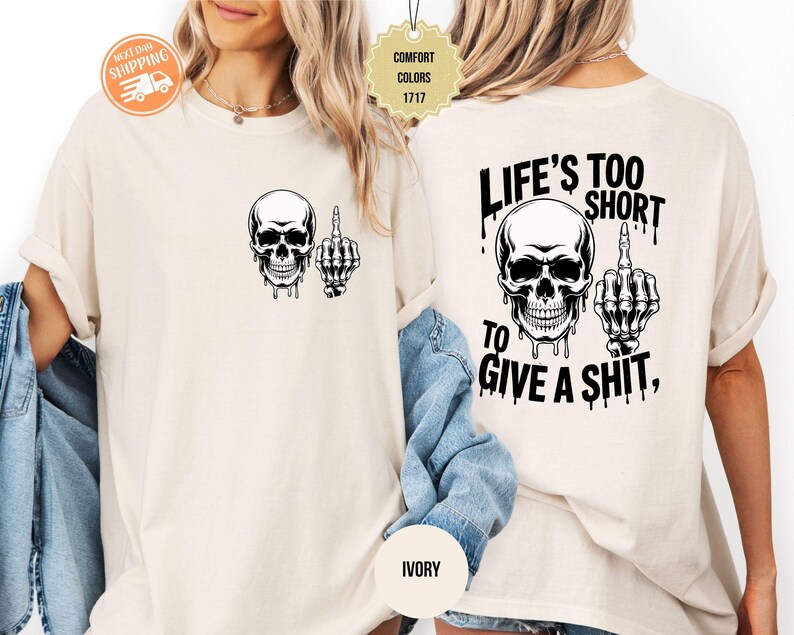 May include: Ivory-colored t-shirt featuring a black skull graphic with a middle finger gesture. The back of the shirt displays the text "LIFE'S TOO SHORT TO GIVE A SHIT," along with a skull and hand graphic. The shirt is a comfort color.