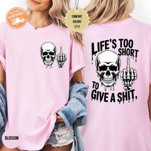 May include: Light pink t-shirt featuring a black and white skull graphic with a middle finger hand gesture. The back of the shirt displays the text "LIFE'S TOO SHORT TO GIVE A SHIT," also in black.