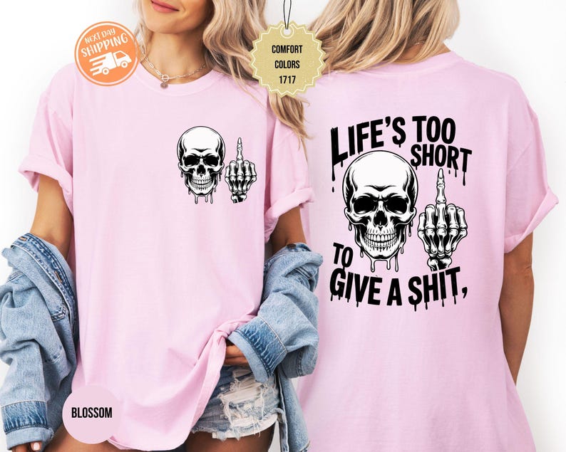May include: Light pink t-shirt featuring a black and white skull graphic with a middle finger hand gesture. The back of the shirt displays the text "LIFE'S TOO SHORT TO GIVE A SHIT," also in black.