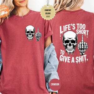 May include: Crimson-colored t-shirt featuring a white skull with a middle finger graphic on the front and back. The back of the shirt has the text "LIFE'S TOO SHORT TO GIVE A SHIT," also with a skull and middle finger graphic.