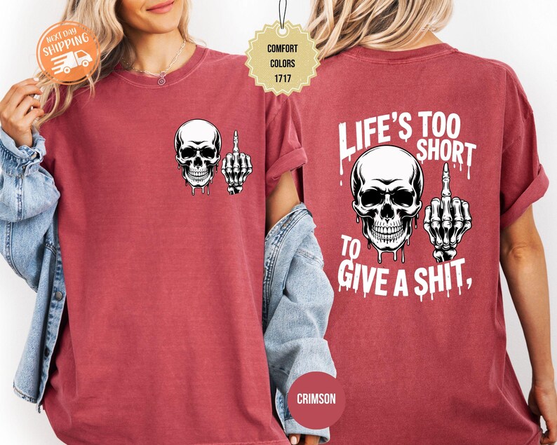 May include: Crimson-colored t-shirt featuring a white skull with a middle finger graphic on the front and back. The back of the shirt has the text "LIFE'S TOO SHORT TO GIVE A SHIT," also with a skull and middle finger graphic.