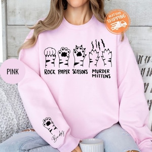 May include: A light pink sweatshirt with a black graphic of cat paws in a "Rock Paper Scissors Murder Mittens" design. A smaller paw print is on the sleeve. The word "Pink" is on a pink circle.