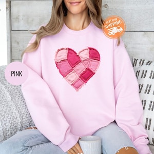 Puede incluir: Una sudadera rosa claro con un diseño de corazón de patchwork en tonos rosas. La palabra "PINK" se muestra en un círculo rosa. La sudadera tiene cuello redondo y mangas largas. Es visible una pegatina de "NEXT DAY SHIPPING".