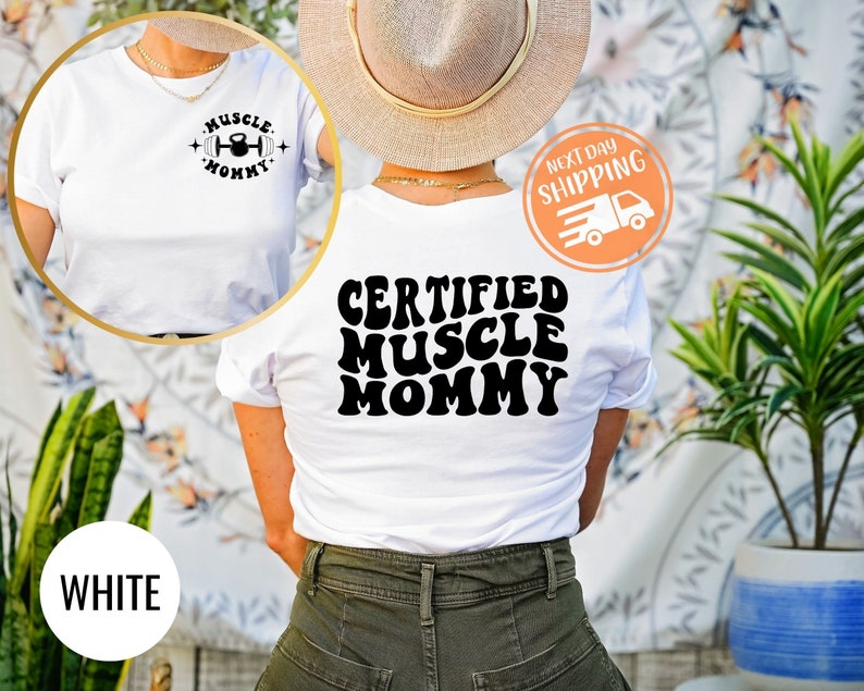 Op de afbeelding: Wit T-shirt met de tekst "Muscle Mommy" in zwart retro-lettertype op de achterkant en een kleine halter- en kettlebell-afbeelding op de voorkant met de tekst "Muscle Mommy" in zwart.
