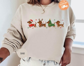 May include: Sand-colored sweatshirt featuring a festive dachshund design. Four dachshunds are dressed in holiday attire: Santa hat, reindeer antlers, string lights, and a Christmas tree. The sweatshirt has a crew neck and long sleeves.