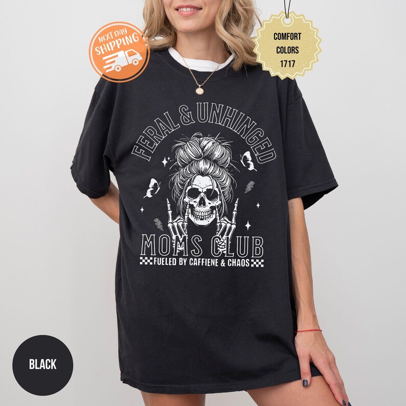 Puede incluir: Camiseta negra con un dise&ntilde;o de calavera y el texto "FERAL & UNHINGED MOMS CLUB". El dise&ntilde;o incluye mariposas y la frase "FUELED BY CAFFIENE & CHAOS". La camiseta es de color confort.