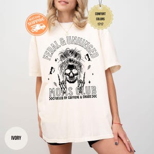 Puede incluir: Camiseta de color marfil con el texto "FERAL & UNHINGED MOMS CLUB" y un gr&aacute;fico de calavera. La camiseta tambi&eacute;n presenta la frase "FUELED BY CAFFIENE & CHAOS". La camiseta es de la marca Comfort Colors.