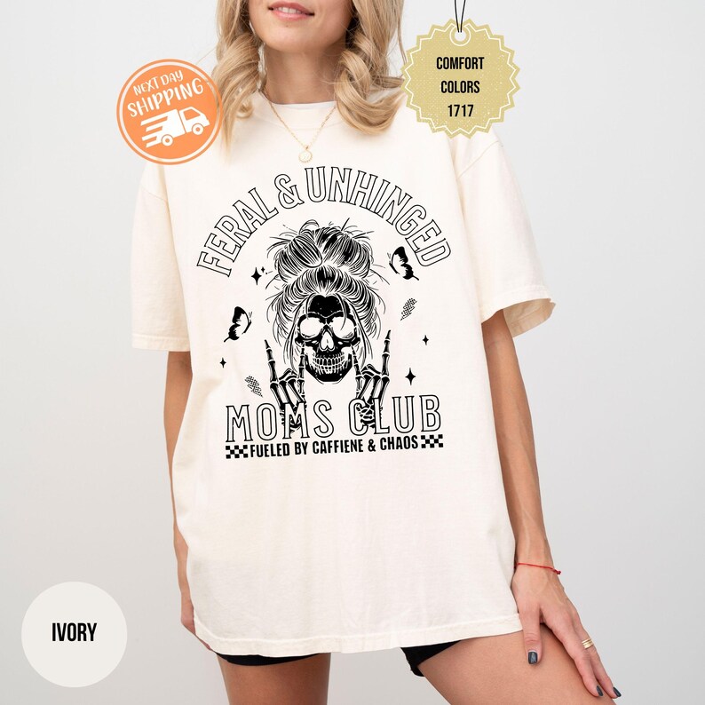 Puede incluir: Camiseta de color marfil con el texto "FERAL & UNHINGED MOMS CLUB" y un gr&aacute;fico de calavera. La camiseta tambi&eacute;n presenta la frase "FUELED BY CAFFIENE & CHAOS". La camiseta es de la marca Comfort Colors.
