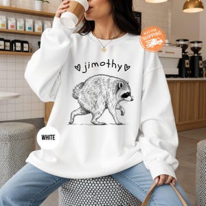 May include: A white crewneck sweatshirt featuring a black ink-style illustration of a raccoon walking, centered above the text "jimothy" flanked by two small heart symbols. The design is printed on the front of the long-sleeved pullover garment.
