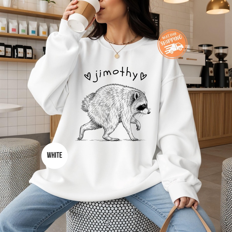 May include: A white crewneck sweatshirt featuring a black ink-style illustration of a raccoon walking, centered above the text "jimothy" flanked by two small heart symbols. The design is printed on the front of the long-sleeved pullover garment.