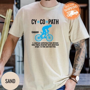 May include: Sand-colored t-shirt with the text "CYCO PATH" above a blue graphic of a cyclist on a bicycle. Below the graphic is the definition of "CYCO PATH". The shirt also has a "Next Day Shipping" graphic.
