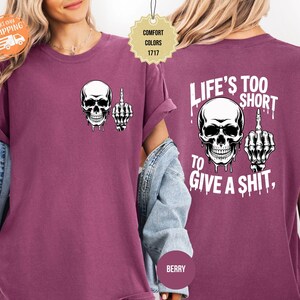 May include: Berry-colored t-shirt featuring a white skull graphic with a raised middle finger on the front. The back of the shirt displays a larger skull graphic and the text "LIFE'S TOO SHORT TO GIVE A SHIT."