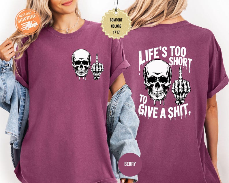 May include: Berry-colored t-shirt featuring a white skull graphic with a raised middle finger on the front. The back of the shirt displays a larger skull graphic and the text "LIFE'S TOO SHORT TO GIVE A SHIT."