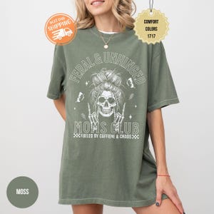Puede incluir: Camiseta verde musgo con el texto "FERAL & UNHINGED MOMS CLUB" y un gr&aacute;fico de calavera. La camiseta tambi&eacute;n incluye la frase "FUELED BY CAFFIENE & CHAOS". La camiseta es de la marca Comfort Colors.