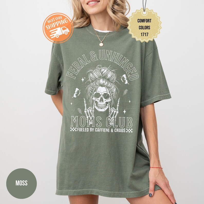Puede incluir: Camiseta verde musgo con el texto "FERAL & UNHINGED MOMS CLUB" y un gr&aacute;fico de calavera. La camiseta tambi&eacute;n incluye la frase "FUELED BY CAFFIENE & CHAOS". La camiseta es de la marca Comfort Colors.