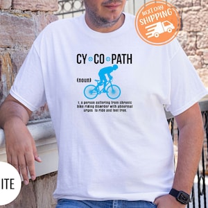 May include: White t-shirt with the text "CY CO PATH" above a blue bicycle graphic. The text below the graphic defines "CY CO PATH" as a person with a chronic bike riding disorder. The shirt also has a "NEXT DAY SHIPPING" sticker.