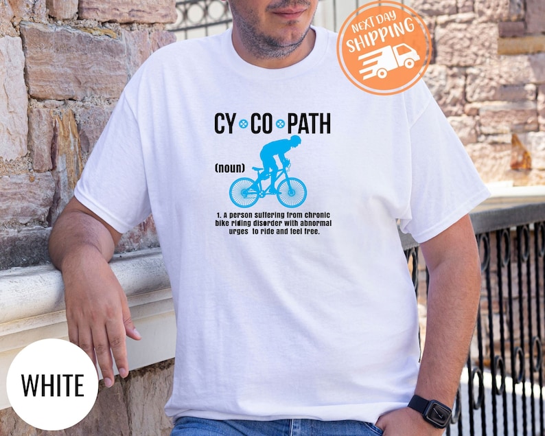 May include: White t-shirt with the text "CY CO PATH" above a blue bicycle graphic. The text below the graphic defines "CY CO PATH" as a person with a chronic bike riding disorder. The shirt also has a "NEXT DAY SHIPPING" sticker.