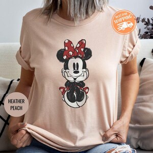 May include: Heather peach-colored t-shirt featuring a Minnie Mouse graphic. The cartoon character is depicted in black, white, and red, with a polka-dot bow. The shirt has rolled-up sleeves and a Next Day Shipping sticker.
