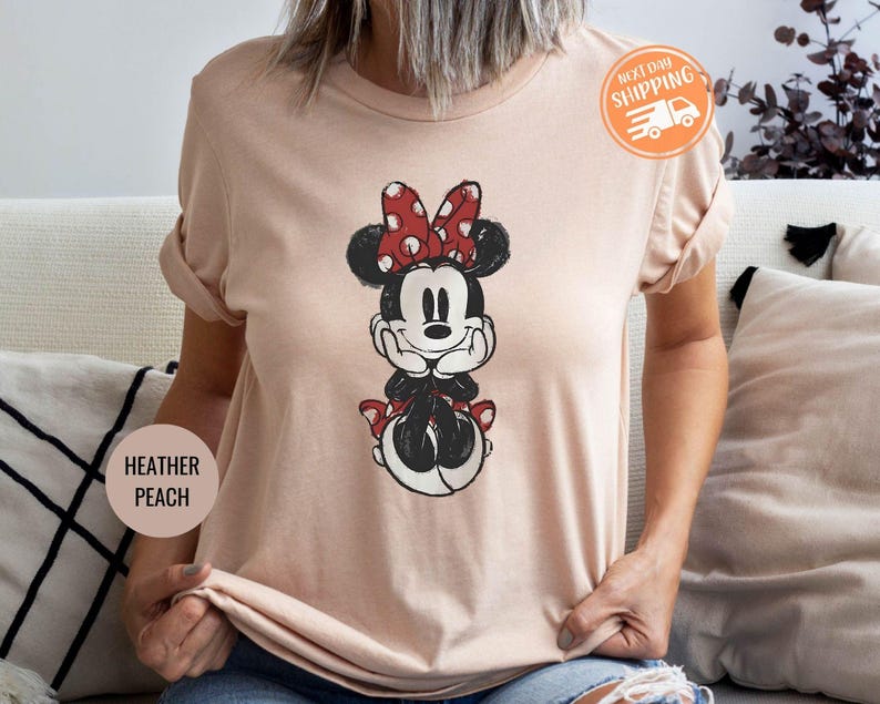 May include: Heather peach-colored t-shirt featuring a Minnie Mouse graphic. The cartoon character is depicted in black, white, and red, with a polka-dot bow. The shirt has rolled-up sleeves and a Next Day Shipping sticker.