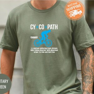 May include: Military green t-shirt with the text "CY CO PATH" in white. A blue graphic of a cyclist is above the definition of "cyclopath". The definition reads: "A person suffering from chronic bike riding disorder with abnormal urges to ride and feel free."