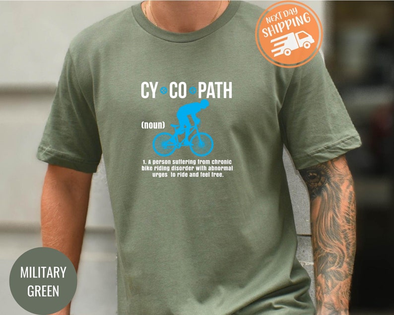 May include: Military green t-shirt with the text "CY CO PATH" in white. A blue graphic of a cyclist is above the definition of "cyclopath". The definition reads: "A person suffering from chronic bike riding disorder with abnormal urges to ride and feel free."