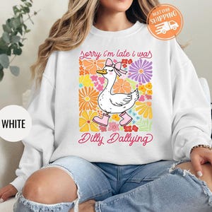 May include: White sweatshirt with a graphic of a cartoon goose wearing pink boots and a pink bow. The text "Sorry I'm late I was dilly dallying" is printed below the goose.