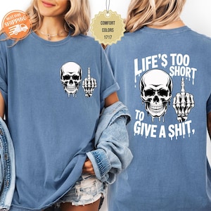 May include: A blue t-shirt featuring a white skull graphic with a raised middle finger. The text "LIFE'S TOO SHORT TO GIVE A SHIT" is printed on the back. The shirt is made of a comfortable material.