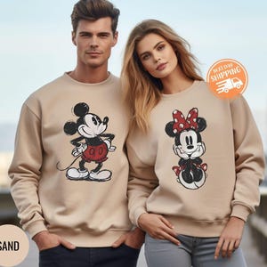 May include: Sand-colored sweatshirts featuring Mickey and Minnie Mouse. Mickey is in a red shorts and Minnie has a red polka dot bow. The sweatshirts are a neutral color and suitable for couples or Disney fans.