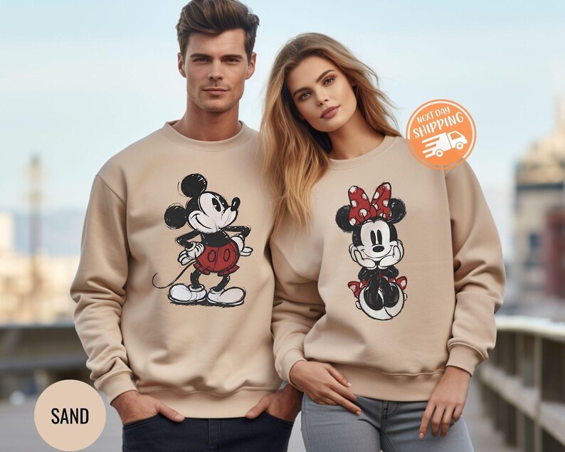 May include: Sand-colored sweatshirts featuring Mickey and Minnie Mouse. Mickey is in a red shorts and Minnie has a red polka dot bow. The sweatshirts are a neutral color and suitable for couples or Disney fans.