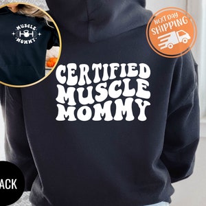 Op de afbeelding: Zwarte hoodie met een witte afbeelding met de tekst "CERTIFIED MUSCLE MOMMY" in een retro-lettertype.