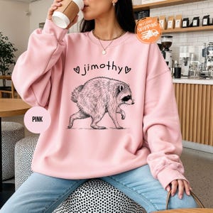 May include: A light pink crewneck sweatshirt featuring a black line-art illustration of a raccoon walking. Above the raccoon, the text reads jimothy, flanked by two small heart outlines. The sweatshirt has long sleeves and a ribbed neckline.