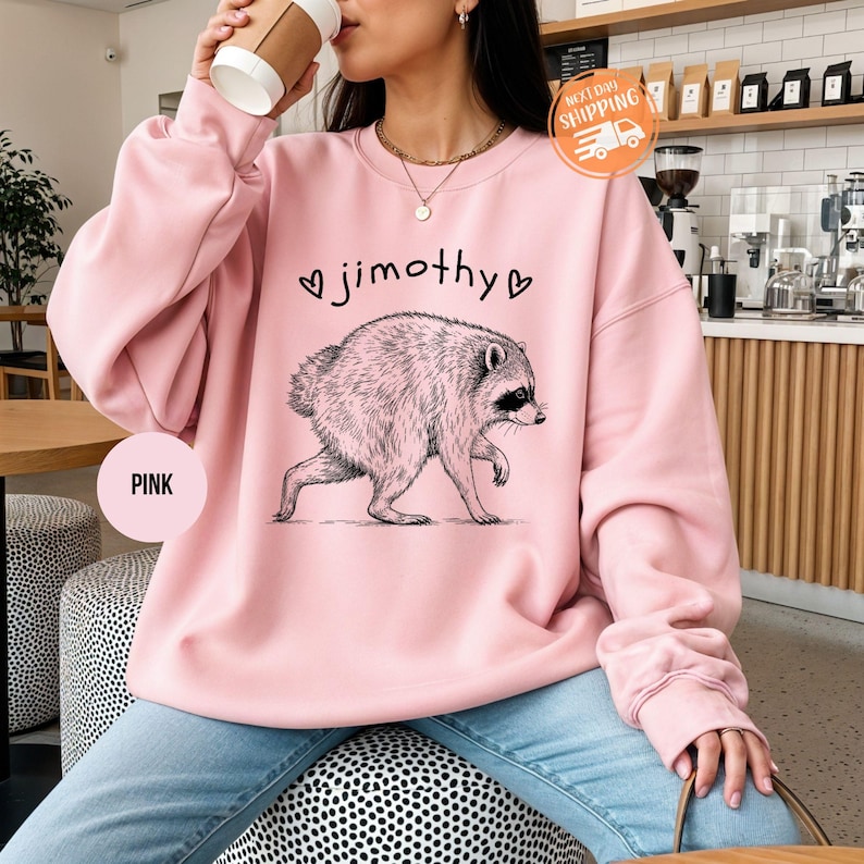 May include: A light pink crewneck sweatshirt featuring a black line-art illustration of a raccoon walking. Above the raccoon, the text reads jimothy, flanked by two small heart outlines. The sweatshirt has long sleeves and a ribbed neckline.