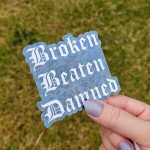 May include: A holographic sticker with the words "Broken Beaten Damned" in white gothic font. The sticker has a blue and silver iridescent effect, and is held by a person with painted nails.