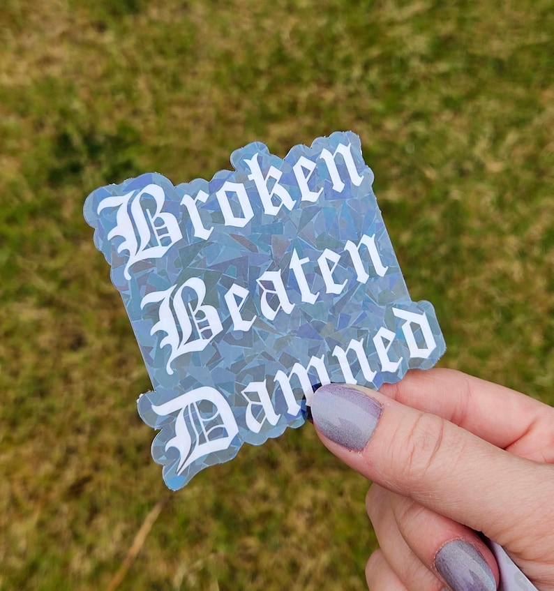 May include: A holographic sticker with the words "Broken Beaten Damned" in white gothic font. The sticker has a blue and silver iridescent effect, and is held by a person with painted nails.