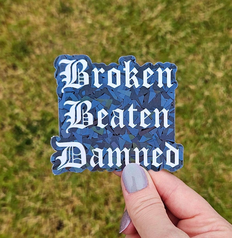 May include: A sticker with the words "Broken Beaten Damned" in a gothic font. The text is white, set against a blue mosaic background. The sticker is held against a backdrop of green grass.