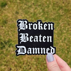 May include: A black sticker with white text that reads "Broken Beaten Damned" in a gothic font. The sticker has a white border and is held against a background of green grass.