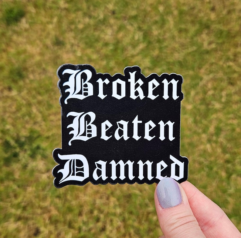 May include: A black sticker with white text that reads "Broken Beaten Damned" in a gothic font. The sticker has a white border and is held against a background of green grass.