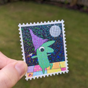 May include: A green cartoon character wearing a purple hat dances on a glittery dance floor with a disco ball in the background. The character is holding a martini glass. The image is printed on a holographic sticker.