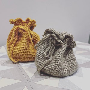 May include: Two crocheted drawstring bags, one in a mustard yellow colour and the other in a light brown colour. The bags are sitting on a tiled surface.