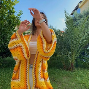 Puede incluir: Un cárdigan de crochet amarillo, naranja y blanco con un corte holgado y mangas largas. El cárdigan está abierto en la parte delantera y tiene un patrón a rayas.