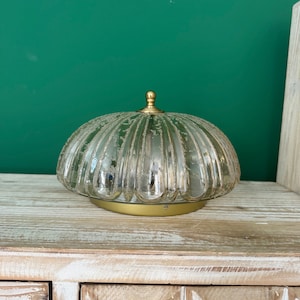 Könnte beinhalten: Eine goldfarbene Lampe mit einem klaren Glasschirm mit strukturiertem, geripptem Design. Der Schirm ist rund und steht auf einer Holzoberfläche.