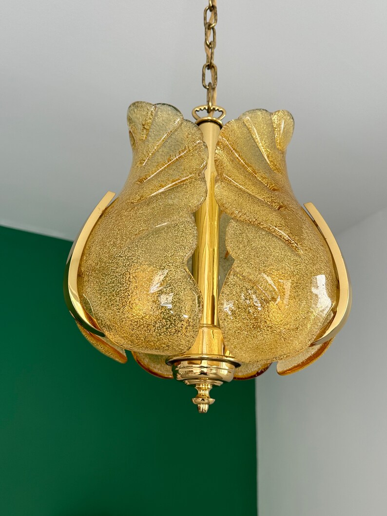 May include: A gold-toned pendant light fixture with a frosted glass shade. The shade is shaped like a flower with multiple petals. The light fixture is hanging from a chain.