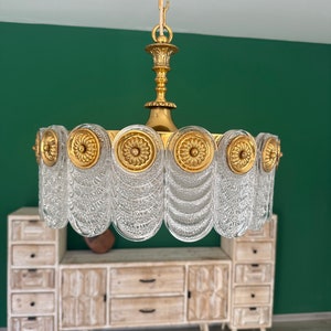 Könnte beinhalten: Ein goldener und durchsichtiger Glas-Kronleuchter mit einem runden Design. Der Kronleuchter hat eine Kette und eine dekorative Spitze.