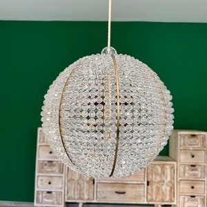 Op de afbeelding: Een ronde, gouden kroonluchter met heldere kristallen kralen. De kroonluchter hangt aan een wit koord tegen een groene muur.