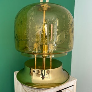 Könnte beinhalten: Eine goldene Tischlampe mit einem grünen Glasschirm. Der Schirm hat ein strukturiertes, blasenartiges Design. Die Lampe steht auf einem Holztisch mit einer Schublade.
