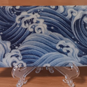 Op de afbeelding: Een rechthoekige keramische tegel met een blauw en wit golfpatroon. De tegel staat op een transparante kunststof standaard.