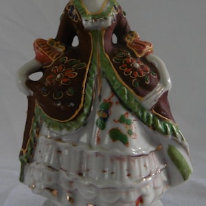 May include: A porcelain figurine of a woman in a period dress. The figure wears a brown coat with floral accents over a white dress. The base and details are trimmed with gold. The woman has a white bow in her hair.