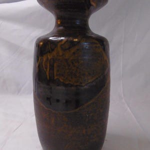 May include: A cylindrical ceramic vase with a narrow neck and a flared rim. The surface features a mottled glaze in shades of dark brown and black, with lighter amber-colored streaks creating a textured, organic appearance across the body.