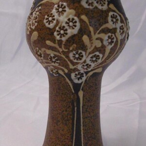 May include: A brown ceramic vase with a textured surface and a flared top. The vase is decorated with a cream-coloured floral design. The vase has a wide base and a slender neck. The overall height is approximately 20 cm.